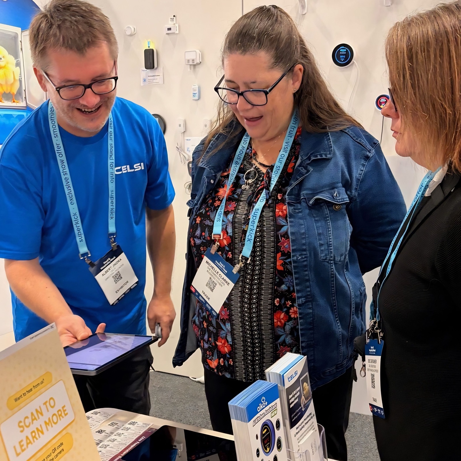 Albert showing two operators the QR food labels at the °Celsi stand, beside a 'Scan to learn more' sign